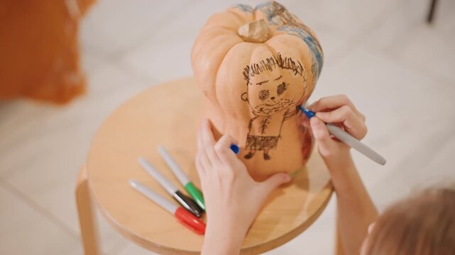 White hands decorating small pumpkin on wooden stool using marker to sketch whimsical face and floral pattern, colored pens arranged on table, warm indoor lighting, cozy autumn craft session, closeup
