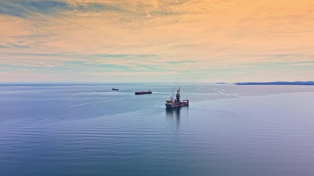 An oil tanker moves through the open sea with another ship visible in the background. The sky shows light colors as the sun begins to rise over the horizon.