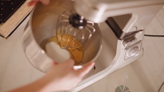 Caucasian hands pouring eggs into mixer bowl with sugar, closeup of stainless steel bowl and whisk attachment, electric appliance on countertop, precise pouring and measuring for pumpkin pie filling,