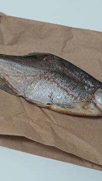 A vertical, slow camera pan captures a single, beautifully dried and salted fish, resembling a vobla or bream, resting on textured brown craft paper against a pristine white surface, emphasizing its
