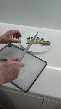 Top-down shot of male hands meticulously slicing a whole dried fish on a clean white cutting board, then artfully assembling the segmented pieces onto a white serving plate, presenting a delicious and