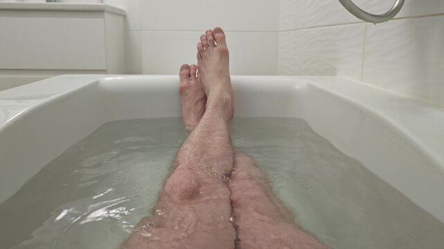 Point-of-view shot of a person's bare legs and feet soaking in a warm, clean bathtub with clear water, enjoying a peaceful moment of relaxation and personal hygiene in a modern white bathroom setting.