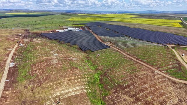 Workers are installing solar panels on a large plot of land. The area is surrounded by green fields, with a blue sky and clouds above. Equipment is present to aid in the installation.