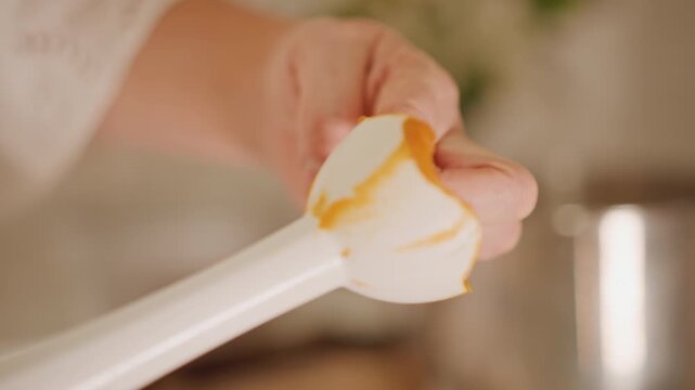 Closeup spatula dipping pumpkin filling into saucepan, hand guiding spatula, glossy orange texture clinging to edge, simmering pot and wooden spoon visible, focused culinary technique