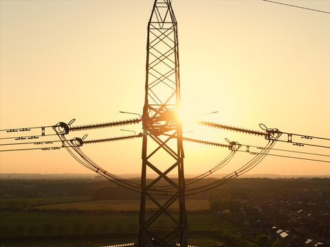 Aerial view of an electricity pylon silhouetted against a golden sunset over green fields and residential houses Armthorpe, England, United Kingdom.