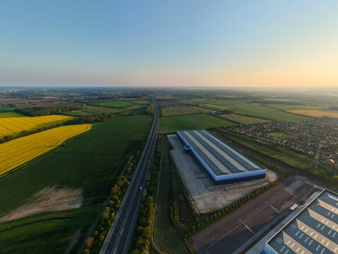 Aerial view of a large modern warehouse facility and highway surrounded by yellow rapeseed fields and residential houses during golden hour in Armthorpe, England, United Kingdom.