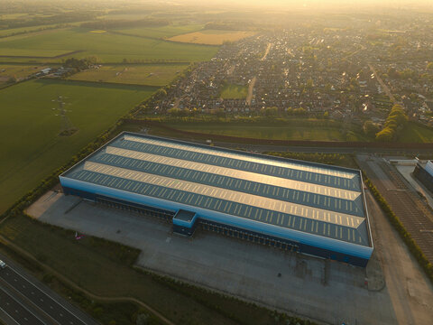 Aerial view of a large industrial warehouse building situated near a residential neighborhood and green fields during golden hour in Armthorpe, England, United Kingdom.