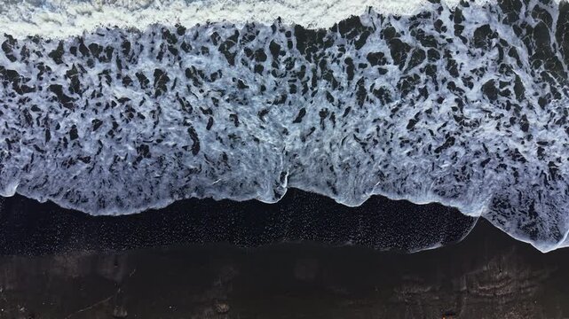Waves break on a black sand beach with a large ocean in view. The water is white and foamy as it meets the shore under a clear sky during the day.