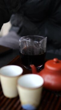 Person performing a traditional chinese tea ceremony, straining hot pu erh tea into a glass pitcher as steam rises from the dark, aromatic liquid, with ceramic cups and a clay teapot nearby