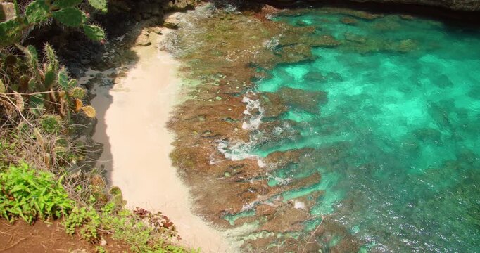 Waves wash over rocky coral tide pools on edge of secluded white sand beach tropical cove