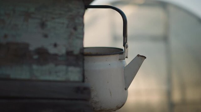 Worn white kettle hanging by shed, muted dusk light with soft silhouette, utilitarian spout and black handle, grime and rust on enamel, greenhouse arc in background, suggests home cook or gardener