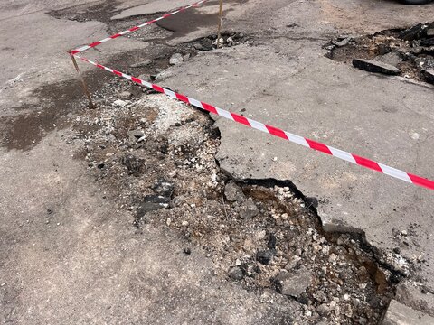 damaged asphalt road with large cracks and a hole, marked with red and white caution tape. Concept of poor infrastructure, road repair, construction work, and public safety