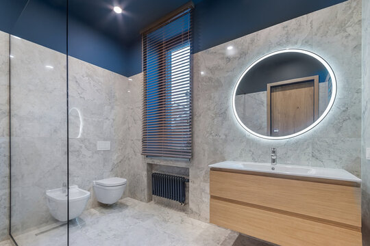 Bathroom with a large round mirror with backlight over the sink and a window with blinds.