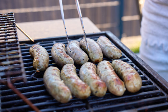 Several sausages cook on barbecue grate while tongs touch one piece