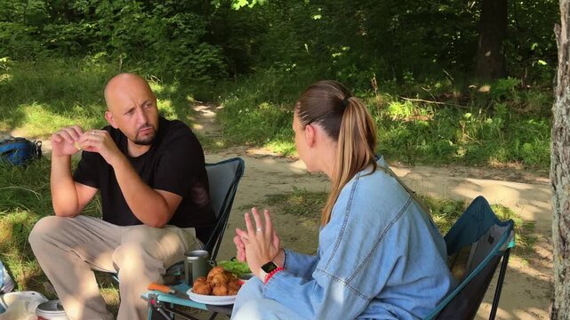 Man and woman enjoying a snack during a camping trip in the forest. Couple sitting in folding chairs and eating sandwiches at a portable table outdoors. Concept of summer picnic and traveler lifestyle