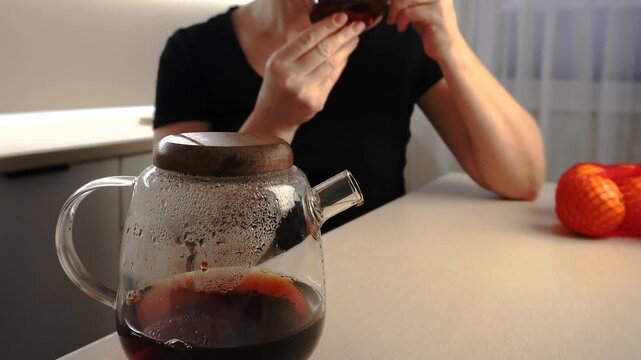 A woman in a black shirt holds a glass cup filled with coffee. A steaming coffee pot sits on the table alongside oranges, creating a cozy atmosphere.