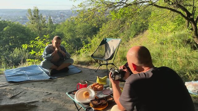 Man with a camera taking a video of a woman meditating on a hill near a campsite. Creative photo and video shooting process in summer nature with a city view background