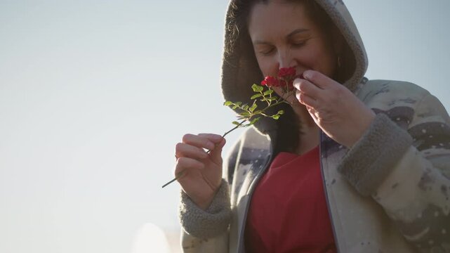 White woman home smelling red rose wearing hooded sweater, tenderly inspecting petals and tucking bloom into cardigan, soft sunlight backlight, suburban backyard, candid closeup, gentle domestic