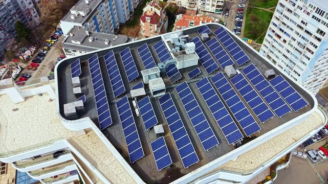 Solar panels are arranged in neat rows on the roof of a large building in a city. Nearby rooftops and cars are visible under clear daylight.