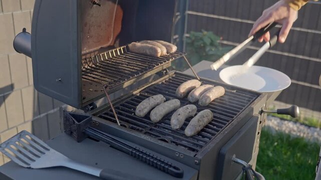 Hand uses tongs to move sausages from upper rack to lower grate on outdoor grill. Barbecue, cooking and backyard food preparation concept