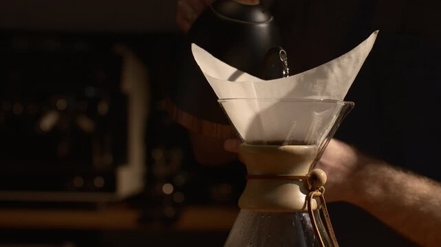 Man preparing filter coffee with Chemex, pouring water in slow motion, dramatic shadows and sunlight, aesthetic specialty coffee process.