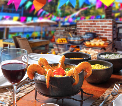 Brazilian shrimp moqueca served in a traditional clay pot, in a festive Festa Junina setting with colorful flags, a wood-burning stove, red wine, and side dishes.