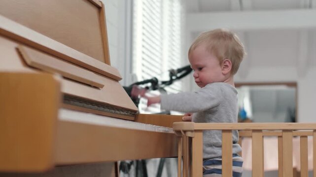 Caucasian toddler plays upright piano in wooden playpen with natural window light, tiny hands pressing keys, playful curiosity and musical discovery