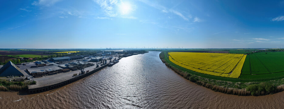 Aerial view of the River Trent flowing past an industrial wharf and vibrant yellow rapeseed fields under a clear blue sky in Amcotts, England, United Kingdom.