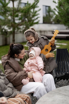 Mother holds baby while older child plays ukulele outside in warm jacket and ear muffs on sunny day