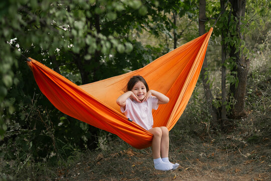Young girl smiles joyfully while sitting in bright orange hammock surrounded by green trees in peaceful forest setting