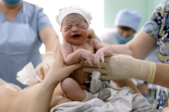 Newborn baby cries loudly held by medical staff in bright hospital room after birth moment