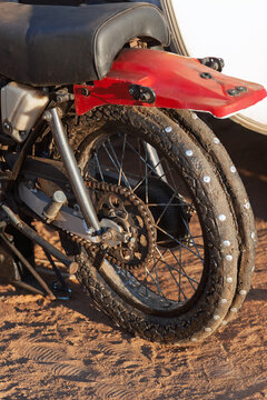 weathered motorcycle tire fitted with metal studs for traction, showing a dirty tread, spokes, and a drive chain on a sandy surface.