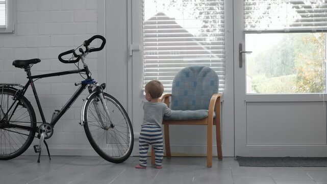 Child explores the space outside. Young kid near bike and chair. Morning light illuminates small child learning to stand. Boy investigates entryway while balancing and reaching for nearby objects
