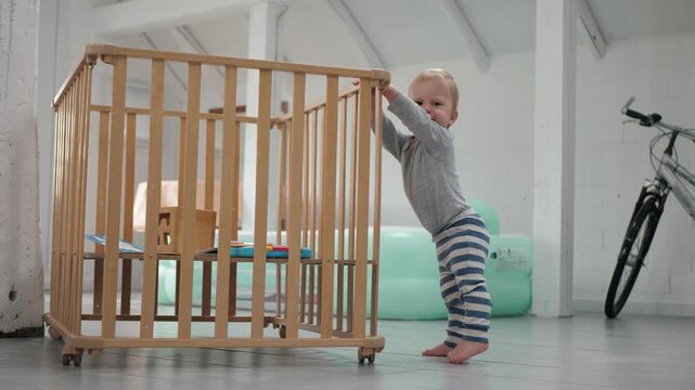 A child explores his surroundings. A toddler in striped pants in a cheerful house. The little explorer carefully examines a wooden playpen