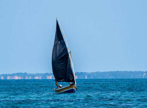 Barca a vela con vela scura inclinata che naviga su mare aperto con orizzonte lontano e cielo limpido, scena marina minimalista con luce naturale.