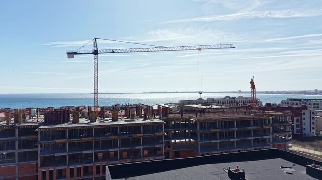 Workers prepare a new building near the ocean. A crane lifts materials for construction under a clear sky. The view shows ongoing development close to the waterfront.