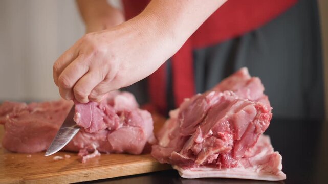 Hands trimming pork on cutting board, white home cook in apron carefully removing fat and silver skin from raw pork, closeup of knife and texture, warm kitchen light, rustic wooden board, domestic