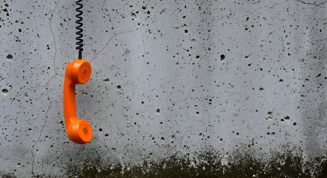 Orange telephone receiver hanging from a black cord against a textured concrete wall with mossy bottom