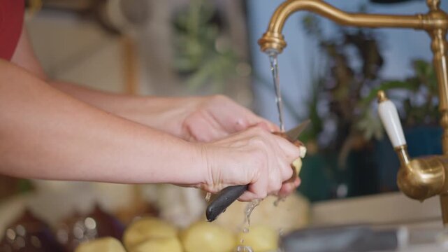 Home cook rinsing potato under tap, water stream and droplets visible, hands turning tuber and cleaning skin, cozy kitchen backdrop with stacked vegetables, focused prep for hearty stew