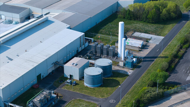 Aerial view of the Industrial Glass Manufacturing Facility with large white warehouses, blue storage tanks, and cooling systems surrounded by green lawns in Goole, England, United Kingdom.