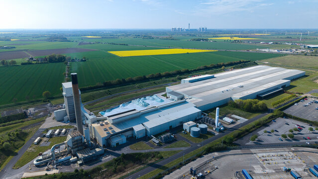 Aerial view of the Industrial Glass Manufacturing Facility with its tall chimney and large warehouses surrounded by green fields and a clear blue sky in Goole, England, United Kingdom.