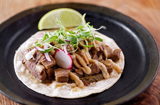 Taco De lengua (beef tongue) on a soft shell tortilla wrap with micro greens and radish with a slice of lime in a dark plate on a wooden table
