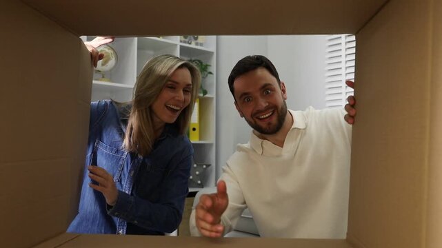 Woman and man opening cardboard box and reaching into it indoors, view from inside package