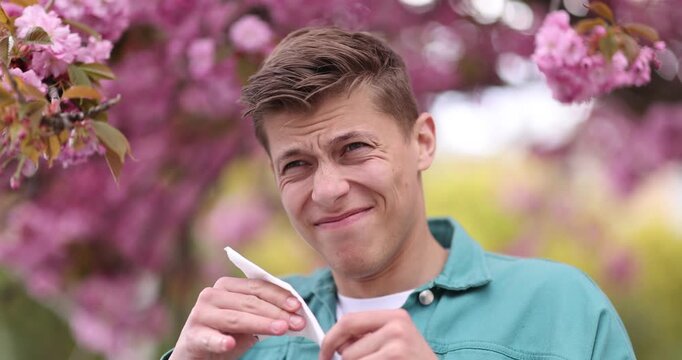 Seasonal allergy. Man with paper tissue sneezing near blossoming tree on spring day