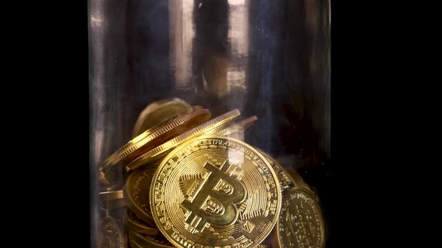 Slow motion footage of golden bitcoin coins falling into glass jar against black background.