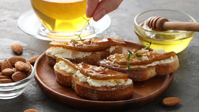 Woman adding thyme to bruschettas with cream cheese, pear and almonds at grey textured table, closeup