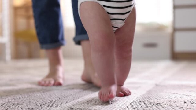 First steps. Mother supporting daughter while she learning to walk at home, closeup