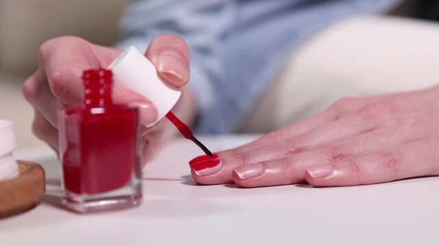 Woman applying red nail polish onto her nails at white table indoors, closeup