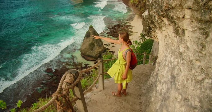 Young woman points at turquoise ocean water from Diamond Beach cliff path in Nusa Penida Bali