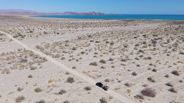 Aerial view shows an SUV driving on dirt roads while towing a teardrop camper trailer close to the ocean in Baja California.
